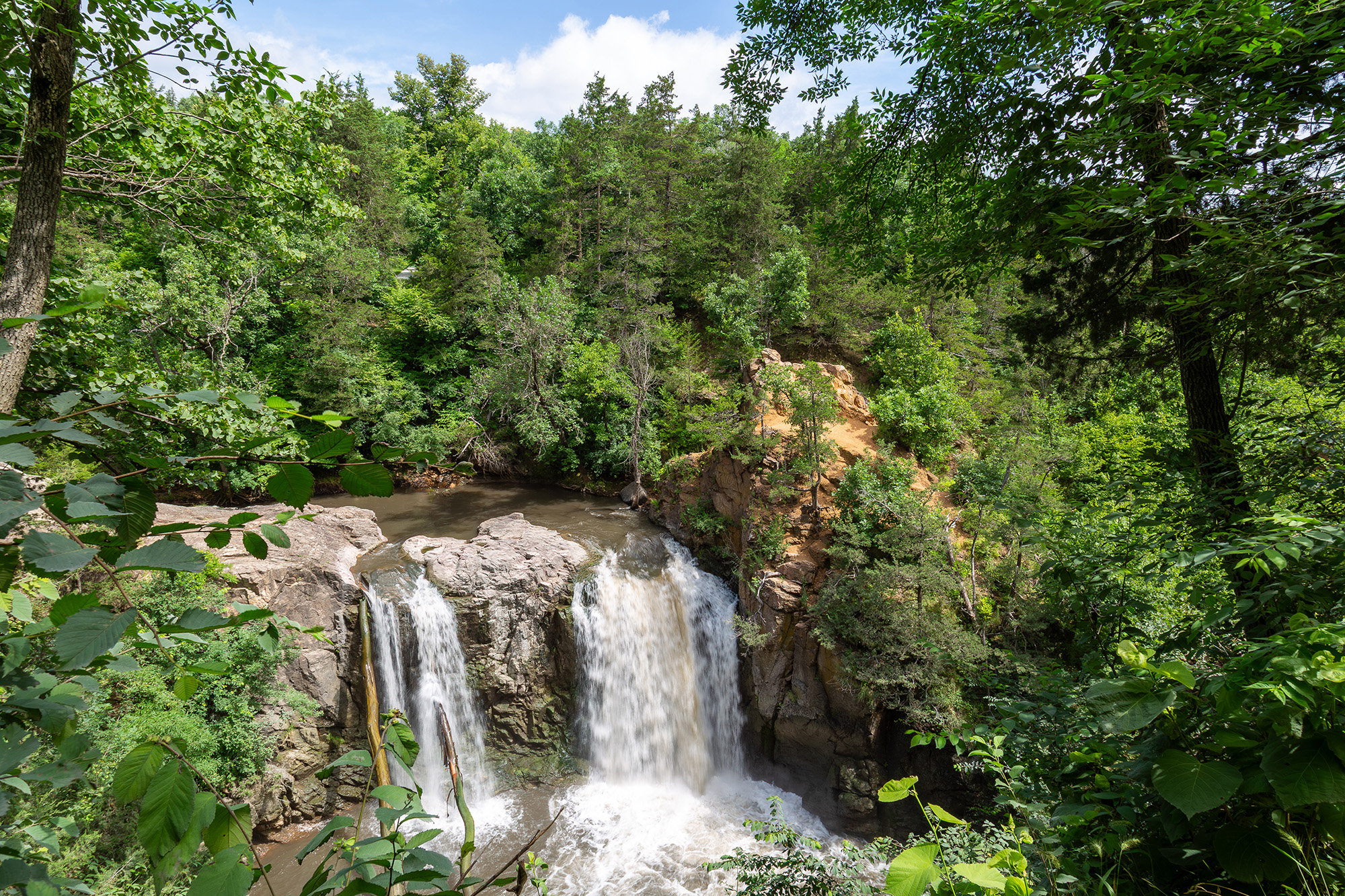 Double split waterfall rushes over the granite rock, surrounded by lush green trees, with a blue sky above.